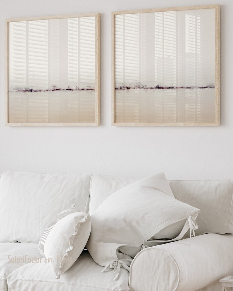 May include: Two framed abstract landscape paintings with a muted color palette of beige, white, and light purple. The paintings are hanging on a white wall above a white couch with white pillows.