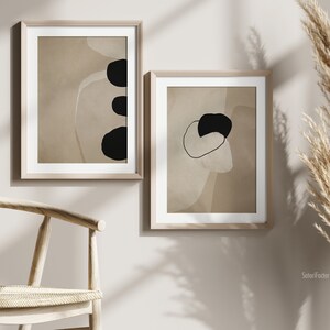 May include: Two framed abstract art prints with black and white shapes on a beige background. The prints are hanging on a light beige wall.