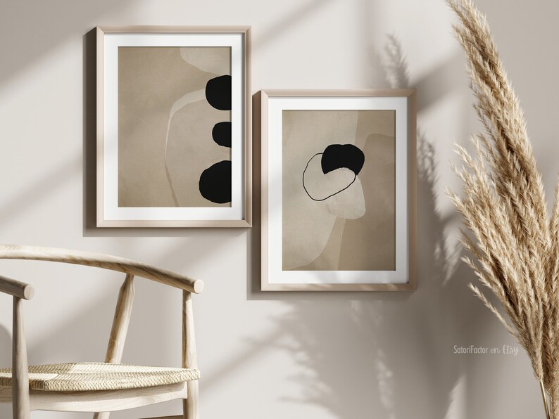 May include: Two framed abstract art prints with black and white shapes on a beige background. The prints are hanging on a light beige wall.