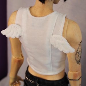 Puede incluir: Un top corto blanco sin mangas con pequeñas alas de tela blanca cosidas en los tirantes. La prenda la lleva una muñeca articulada con tatuajes visibles en los brazos, combinada con pantalones vaqueros negros que incluyen un detalle de cadena plateada.