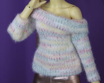Puede incluir: Una muñeca con un suéter blanco de hombros descubiertos con rayas rosas, verdes y amarillas y leggings metálicos dorados.
