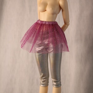 Può includere: Una bambola indossa una gonna tutù in tulle color magenta glitterato sopra dei leggings capri color argento metallizzato. La gonna presenta una trama a rete fine con sottili accenti scintillanti, mentre i leggings hanno una finitura liscia e riflettente.