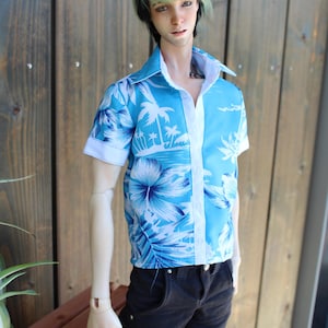 May include: A doll in a short-sleeved shirt featuring a tropical print in blue and white, with a white collar and cuffs. The doll is also wearing black jeans. The shirt's design includes palm trees and floral patterns.