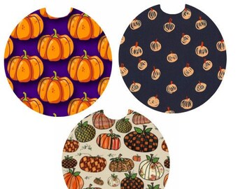 May include: Set of three car coasters with pumpkin designs. One coaster has orange pumpkins on a purple background. Another has sketched pumpkins on a navy background. The third has various pumpkin patterns on a cream background.