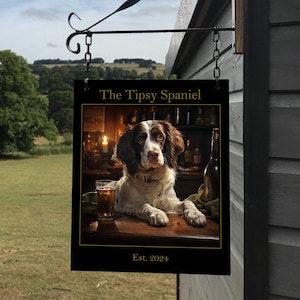 Könnte beinhalten: Ein schwarzes Schild mit goldenen Buchstaben hängt an einer Metallhalterung. Das Schild trägt die Aufschrift "The Tipsy Spaniel Est. 2024". Das Schild zeigt ein Foto eines braunen und weißen Spaniel-Hundes, der an einer Bar mit einem Glas Bier sitzt.