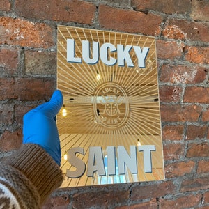 Op de afbeelding: Een gouden spiegel met een zonnestralenontwerp en de woorden "Lucky Saint" in witte letters. De spiegel heeft een klein logo in het midden met een lieveheersbeestje en de woorden "Lucky Saint".