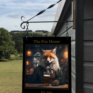 Puede incluir: Un letrero negro con letras doradas cuelga de un soporte de metal negro. El letrero dice "The Fox House Est. 2024". Una ilustración de un zorro con una chaqueta verde y sosteniendo un vaso de cerveza está en el centro del letrero.