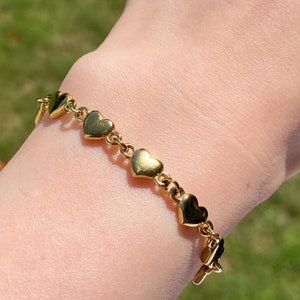 K&ouml;nnte beinhalten: Ein goldenes Armband mit herzf&ouml;rmigen Gliedern und einem Verschluss. Das Armband wird am Handgelenk getragen.