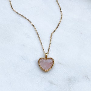 May include: A gold chain necklace with a pink heart-shaped pendant. The pendant has a gold border and a delicate design.