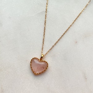May include: A gold chain necklace with a pink heart-shaped pendant. The pendant has a gold border and a delicate design.