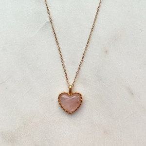 May include: A gold chain necklace with a pink heart-shaped pendant. The pendant has a gold border.
