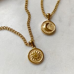 May include: Two gold necklaces with pendants. One pendant features a sun design, the other a crescent moon design. Both pendants are circular and have a textured surface.