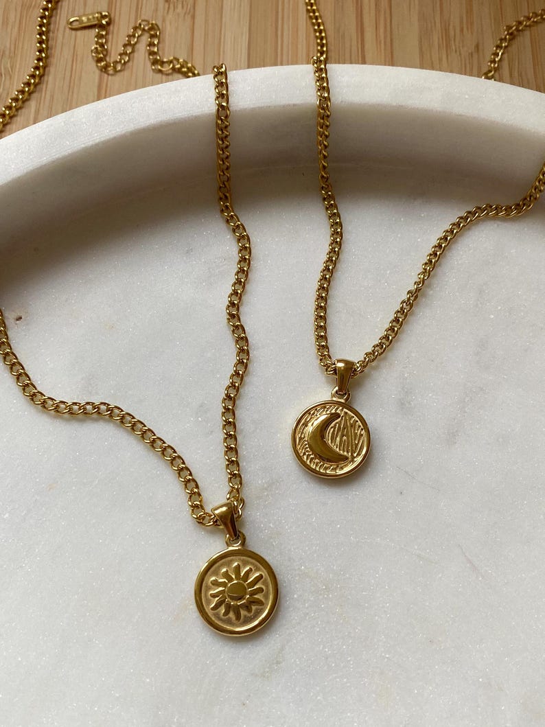 May include: Two gold chain necklaces with pendants. One pendant is a sun with a textured surface and the other is a crescent moon with a textured surface.