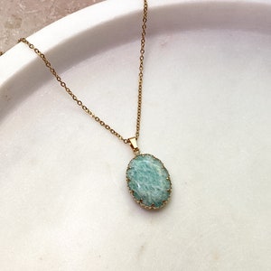 May include: A gold-colored chain necklace with a light blue oval pendant. The pendant has a textured surface and is framed by a decorative gold-colored border. The necklace is displayed on a white marble surface.