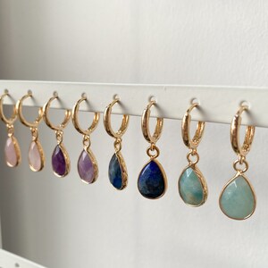 May include: Eight pairs of gold hoop earrings with a teardrop-shaped gemstone pendant. The gemstones are pink, purple, light purple, blue, and green.