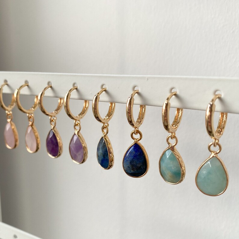 May include: Eight pairs of gold hoop earrings with a teardrop-shaped gemstone pendant. The gemstones are pink, purple, light purple, blue, and green.