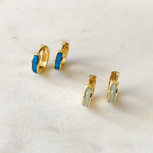 May include: Two pairs of gold hoop earrings with a thin band of opal stones. One pair has blue opal stones and the other pair has white opal stones.