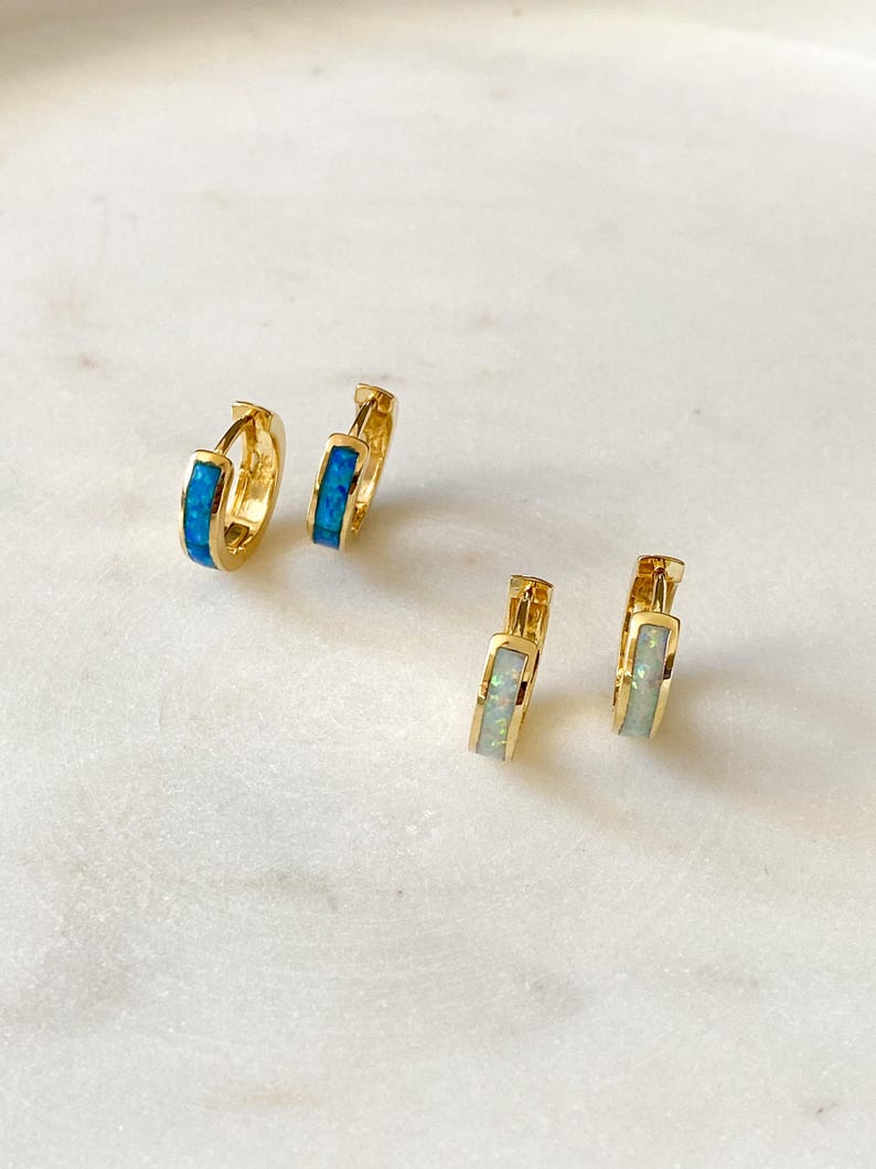 May include: Two pairs of gold hoop earrings with a thin band of opal stones. One pair has blue opal stones and the other pair has white opal stones.
