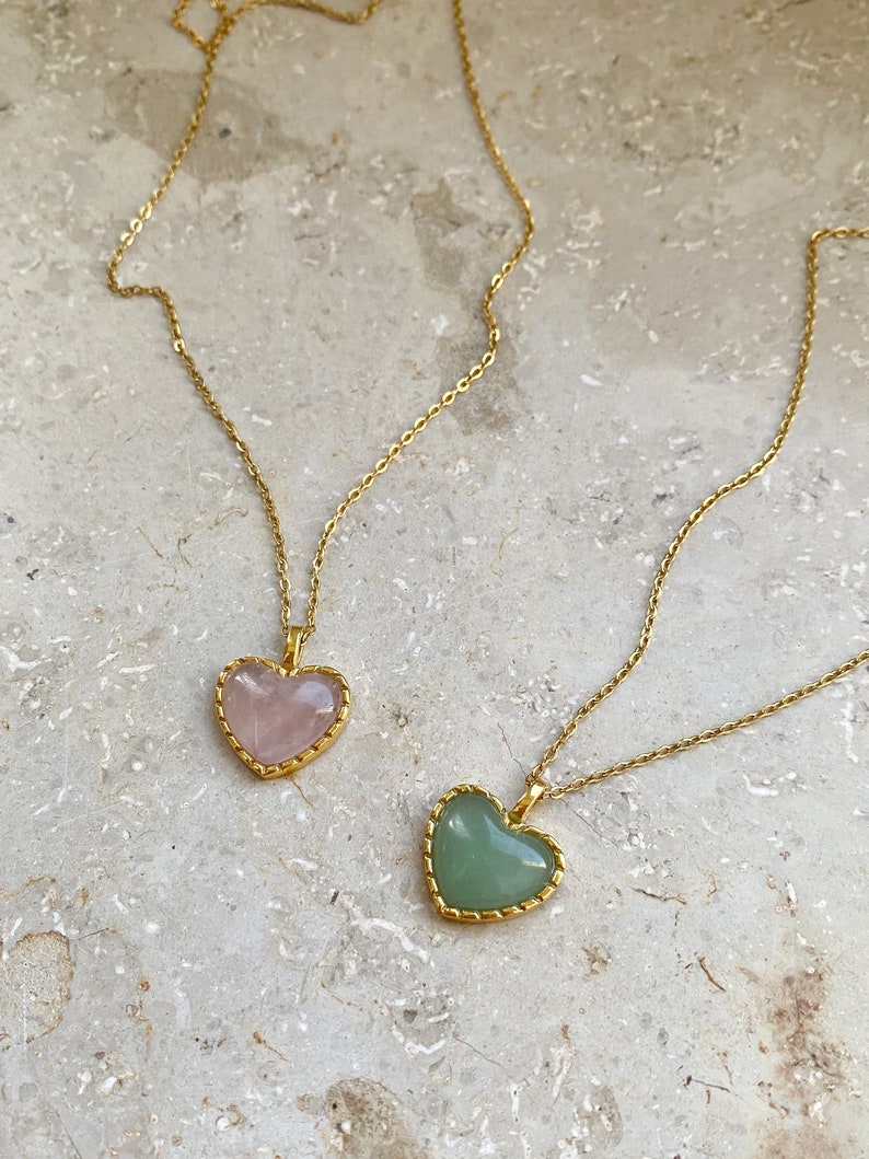 May include: Two gold necklaces with heart-shaped pendants. One pendant is a light pink heart, the other is a light green heart.
