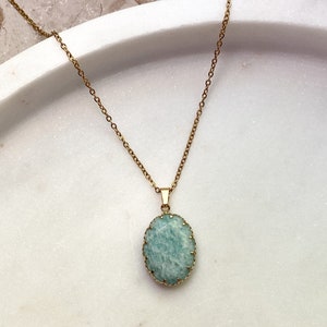 May include: A gold-colored chain necklace with a pendant. The pendant features a light blue oval-shaped stone with a gold-colored bezel setting. The necklace is displayed on a white surface.