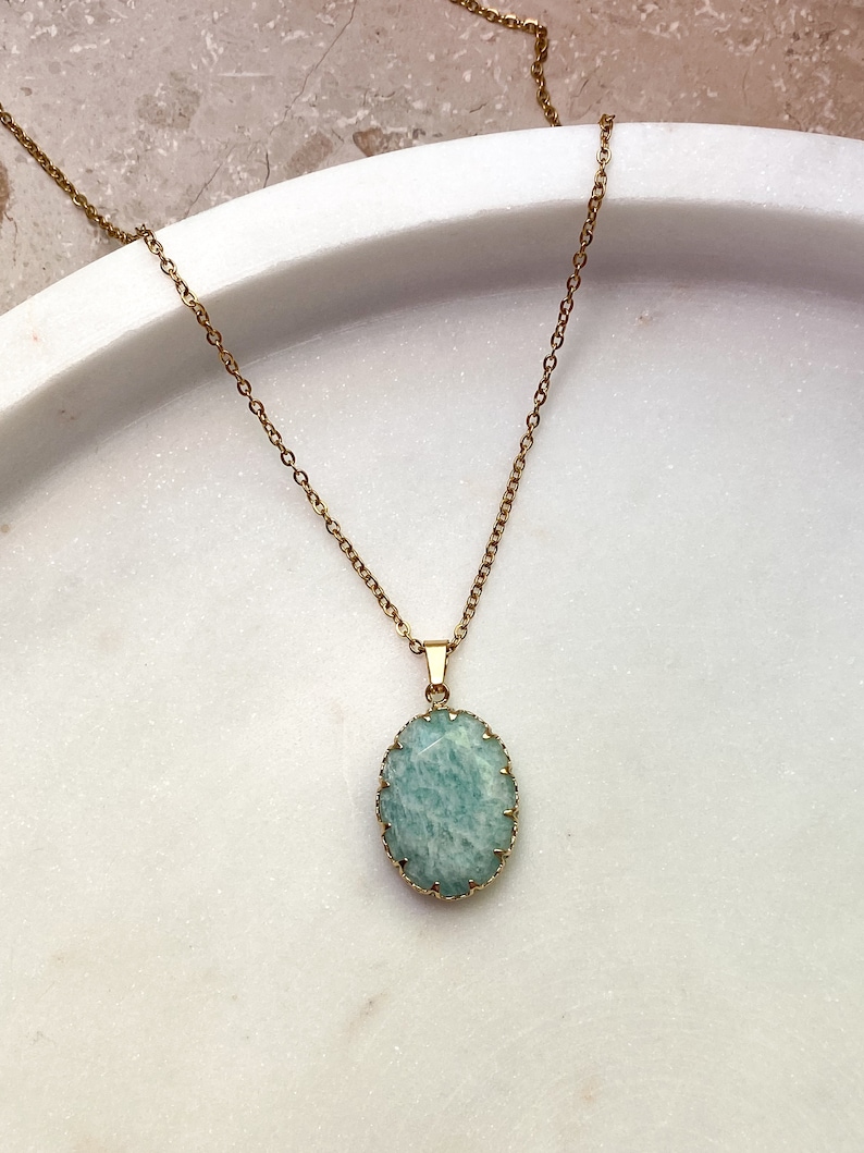 May include: A gold-colored chain necklace with a pendant. The pendant features a light blue oval-shaped stone with a gold-colored bezel setting. The necklace is displayed on a white surface.