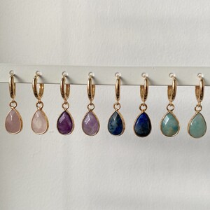 May include: Six pairs of gold hoop earrings with gemstone drops. The gemstones are pink, purple, amethyst, blue, and green.