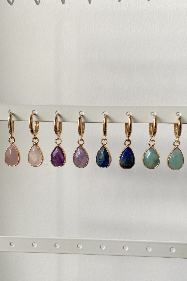May include: Six pairs of gold hoop earrings with gemstone drops. The gemstones are pink, purple, amethyst, blue, and green.