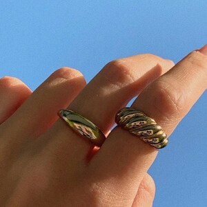 Op de afbeelding: Twee gouden ringen aan een hand. De ene ring is een eenvoudige, gladde band. De andere ring is een dikkere band met een touwachtig ontwerp.