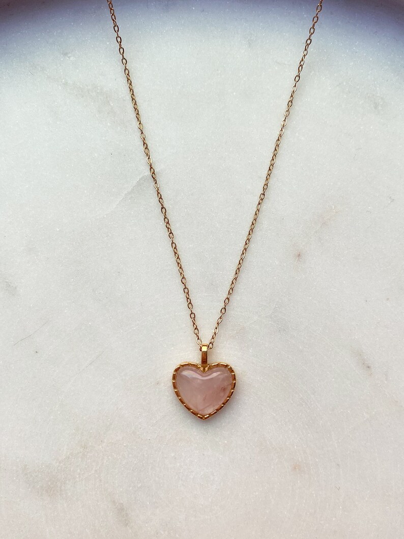 May include: A gold chain necklace with a pink heart-shaped pendant. The pendant has a gold border.