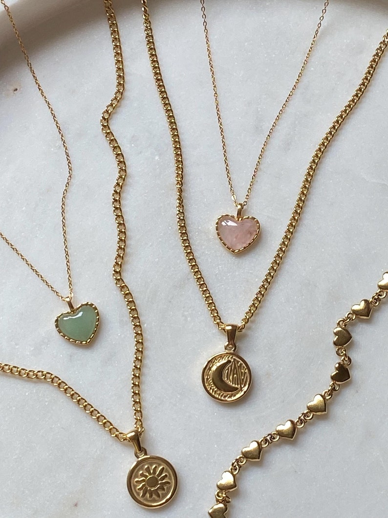 May include: Four gold necklaces with different pendants. One necklace has a green heart-shaped stone pendant, another has a pink heart-shaped stone pendant, another has a gold pendant with a crescent moon and a star, and the last has a gold pendant with a sun. A gold chain with small heart-shaped links is also visible.
