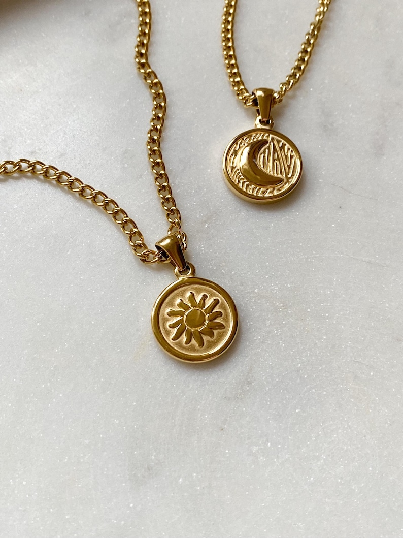 May include: Two gold necklaces with pendants. One pendant features a sun design, the other a crescent moon design. Both pendants are circular and have a textured surface.