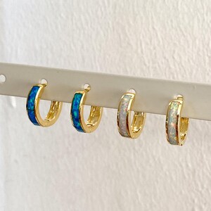 May include: Three pairs of gold hoop earrings with a thin band of opal inlay. The earrings are displayed on a white background. The opal inlay is blue, white, and white.