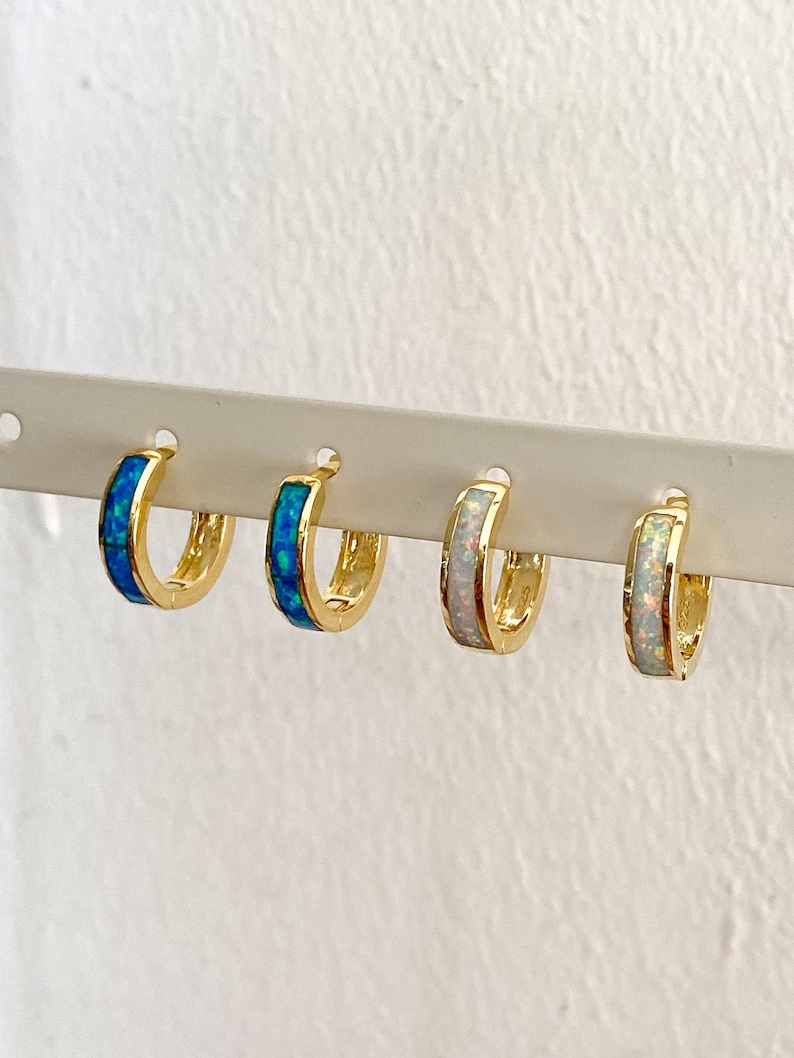 May include: Three pairs of gold hoop earrings with a thin band of opal inlay. The earrings are displayed on a white background. The opal inlay is blue, white, and white.
