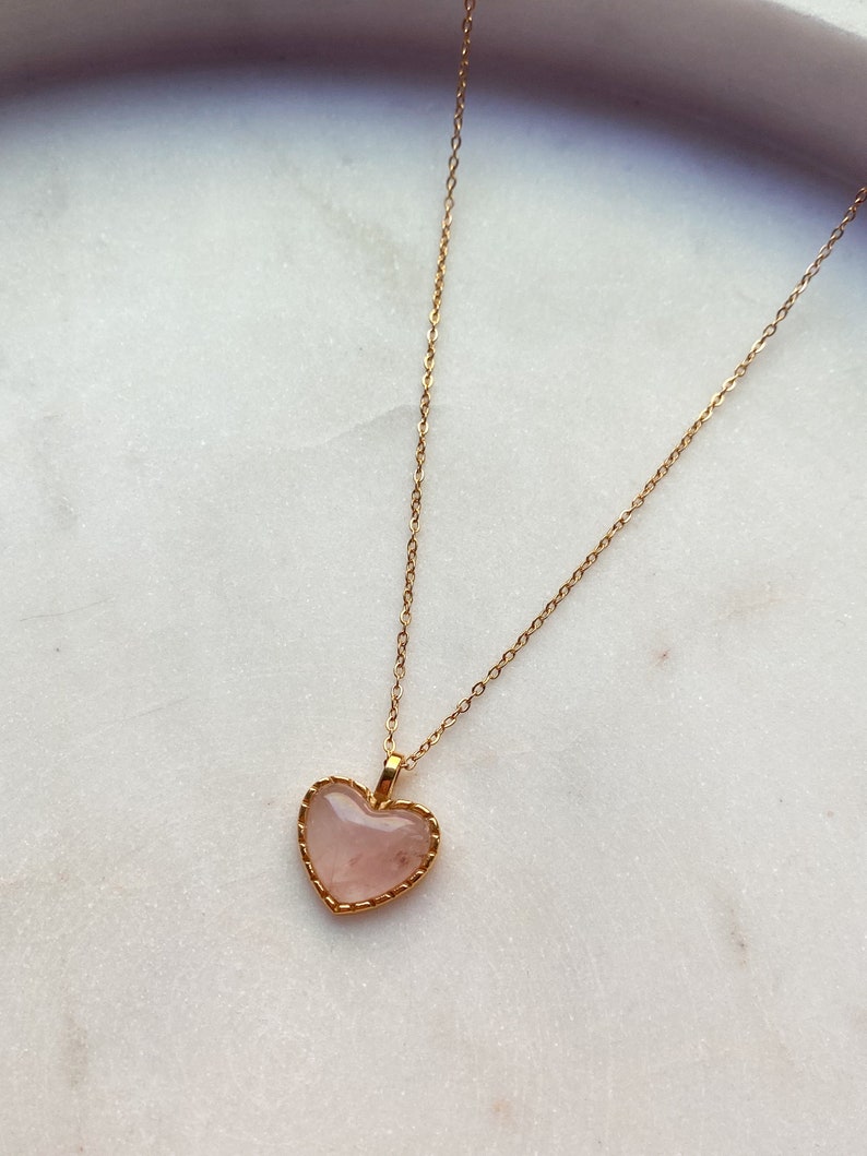 May include: A gold chain necklace with a pink heart-shaped pendant. The pendant has a gold border and a delicate design.