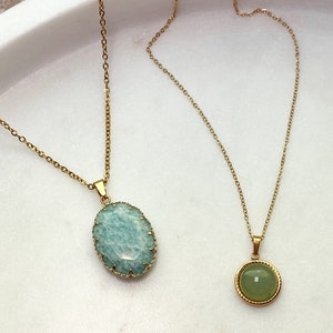 May include: Two gold-toned necklaces with gemstone pendants. One features a light blue, oval-shaped stone with a textured gold frame. The other has a small, round, green stone with a gold border. Both hang on delicate gold chains.