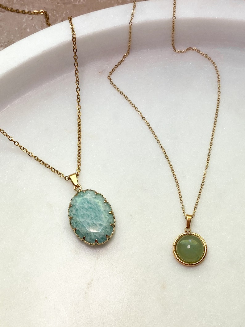 May include: Two gold-toned necklaces with gemstone pendants. One features a light blue, oval-shaped stone with a textured gold frame. The other has a small, round, green stone with a gold border. Both hang on delicate gold chains.