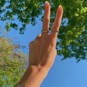 Op de afbeelding: Een hand met twee gouden ringen aan de vingers, die een peace-teken maakt. De hand draagt een gouden horloge met een vierkante wijzerplaat. De achtergrond is een blauwe lucht met groene bomen.