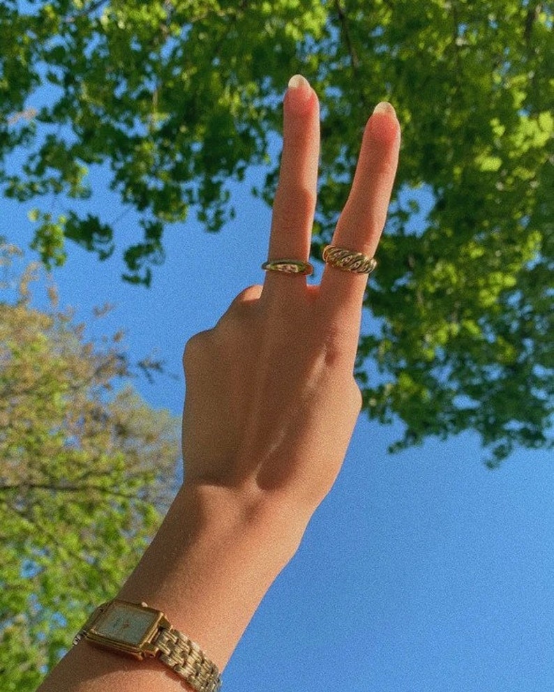 Op de afbeelding: Een hand met twee gouden ringen aan de vingers, die een peace-teken maakt. De hand draagt een gouden horloge met een vierkante wijzerplaat. De achtergrond is een blauwe lucht met groene bomen.
