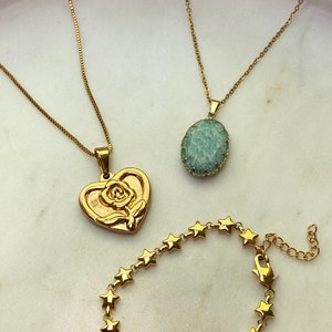 May include: Gold-toned jewelry set on a white surface. Includes two necklaces, one with a heart pendant featuring a rose, and another with a light green oval stone. A star-shaped bracelet is also present.