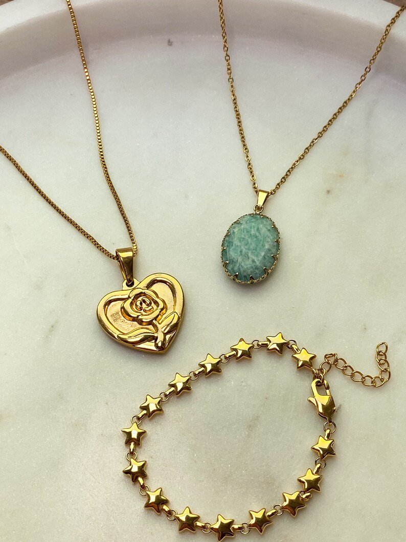 May include: Gold-toned jewelry set on a white surface. Includes two necklaces, one with a heart pendant featuring a rose, and another with a light green oval stone. A star-shaped bracelet is also present.