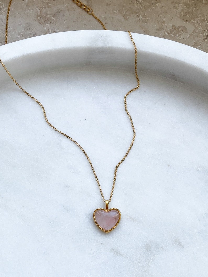 May include: A gold chain necklace with a pink heart-shaped pendant. The pendant has a gold border and a delicate design.