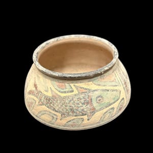 May include: A small, round, beige ceramic bowl featuring a hand-painted design of a fish in shades of green, red, and black. The bowl has a wide, open top with a dark, weathered rim and a textured, matte surface finish throughout the exterior.