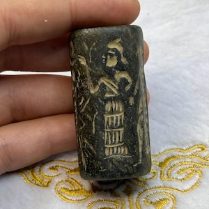 Könnte beinhalten: Ein dunkelgrüner Zylinderstempel mit einem geschnitzten Design einer Figur in einem langen Gewand und einer Kopfbedeckung. Die Figur hält einen Stab und ist von anderen Figuren und Symbolen umgeben.
