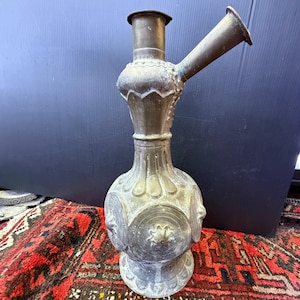 May include: An antique metal hookah with a bulbous base and a long, angled spout. The hookah has intricate decorative details and a tarnished finish, suggesting age and use. The piece sits on a patterned rug.