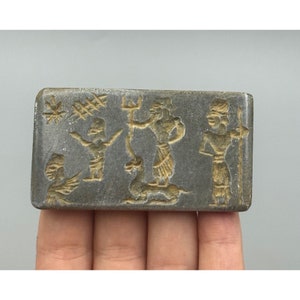 May include: A rectangular grey stone seal with a carved design of three figures. The figures are stylized and appear to be wearing clothing. The seal is approximately 2 inches long and 1 inch wide.