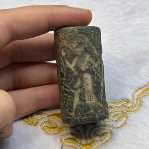Könnte beinhalten: Ein grauer Steinzylindersiegel mit einer geschnitzten Abbildung einer Figur, die einen Stab hält. Die Figur ist von komplizierten Details umgeben.