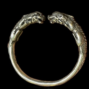 Puede incluir: Pulsera de plata con dos cabezas de dragón que se miran. La pulsera está hecha de metal liso y pulido y tiene un diseño sencillo.