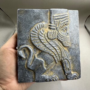 May include: A gray stone relief sculpture of a winged lion with a human head. The lion is depicted in profile, with its head turned to the right. The lion's wings are spread out behind it, and its tail is curled around its legs. The lion's face is detailed, with a beard and a crown.