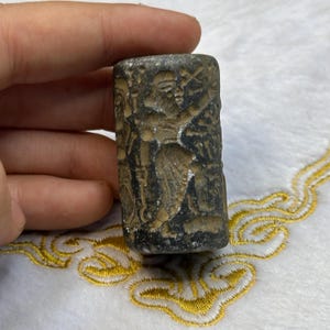 Könnte beinhalten: Ein zylindrisches Steingedicht in dunkelgrau mit aufwendigen Schnitzereien einer Figur und anderer Symbole. Das Siegel ist etwa 5 cm lang und 2,5 cm im Durchmesser.