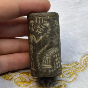 Könnte beinhalten: Ein dunkelgrauer zylindrischer Steinstempel mit aufwendigen Schnitzereien von zwei Figuren und Symbolen. Der Stempel ist mit einem detaillierten Design von zwei Figuren geschnitzt, eine steht, die andere sitzt, mit einem Symbol über ihnen. Der Stempel ist ein historisches Artefakt.